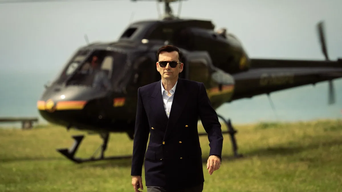 David Tennant as Tony, Lord Baddingham, walking away from a helicopter wearing sunglasses