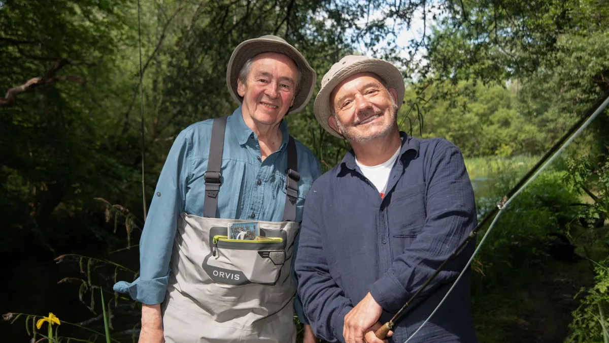 Paul and Bob pose side by side alongside a river with Bob holding a fishing rod