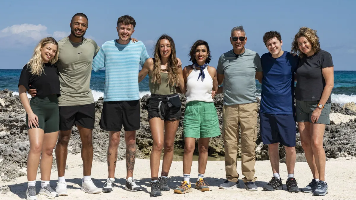 Cast of the show pose side by side for a group photo on a sandy beach